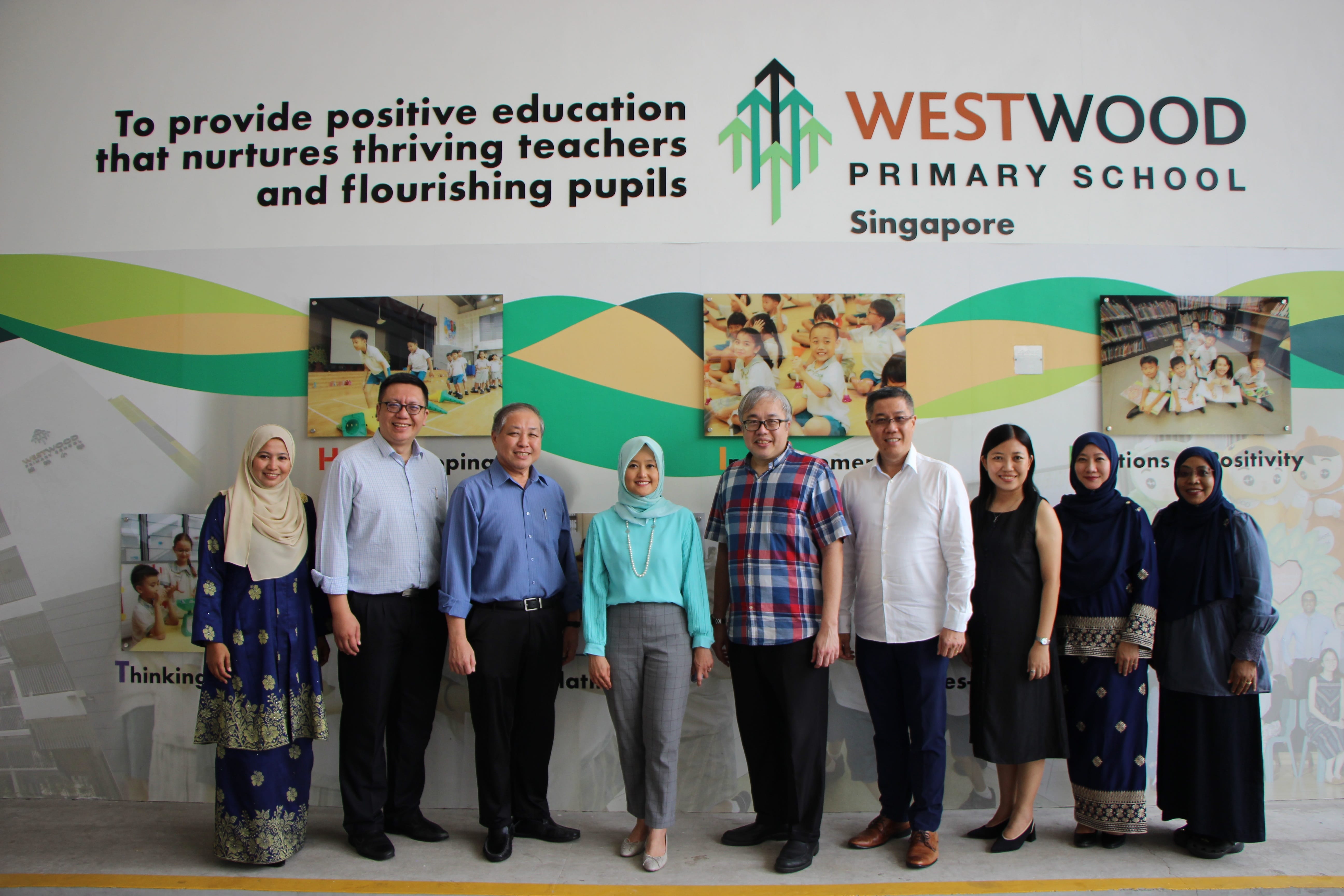 Eight adults stand posing in front of a Westwood Primary School Singapore wall display.